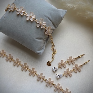 May include: A gold necklace, bracelet, and earrings set with delicate flower charms and clear stones. The jewelry is on a light gray surface.