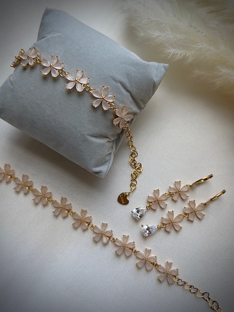 May include: A gold necklace, bracelet, and earrings set with delicate flower charms and clear stones. The jewelry is on a light gray surface.