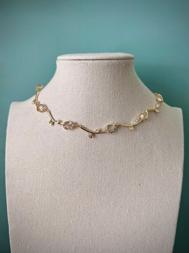 May include: A gold necklace with a delicate vine design and clear stones. The necklace is displayed on a white mannequin.