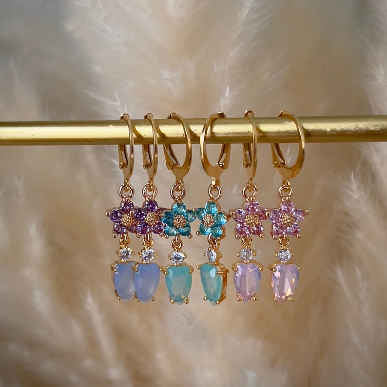 Puede incluir: Cinco pares de pendientes colgantes de oro con un dise&ntilde;o de flor y un colgante en forma de coraz&oacute;n. Las flores est&aacute;n hechas de piedras de cristal de colores y los colgantes son de vidrio opalescente.