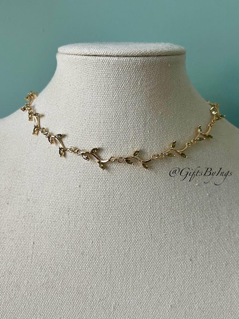 May include: Gold necklace with a delicate vine design featuring small leaves. The necklace is on a white mannequin bust. The text "@GiftsByLugs" is visible in the bottom right corner of the image.