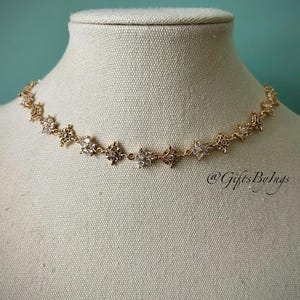 Puede incluir: Un collar de oro con una cadena delicada y pequeños abalorios en forma de flor brillantes. El collar se muestra en un maniquí de busto blanco.
