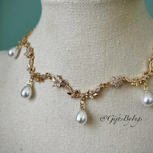 May include: A gold-toned necklace with a floral and leaf design, adorned with clear crystals and dangling teardrop-shaped pearls. The necklace is displayed on a mannequin, showcasing its delicate details and elegant style.