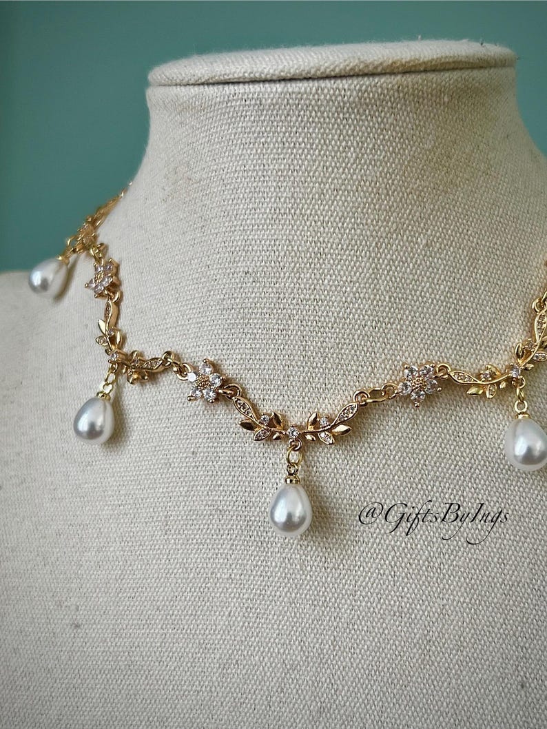 May include: A gold-toned necklace with a floral and leaf design, adorned with clear crystals and dangling teardrop-shaped pearls. The necklace is displayed on a mannequin, showcasing its delicate details and elegant style.