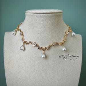 May include: A gold-toned necklace with floral and leaf detailing, adorned with clear crystals and dangling teardrop-shaped pearls. The necklace is displayed on a mannequin head against a teal background. The text "@GiftsByIngs" is visible.