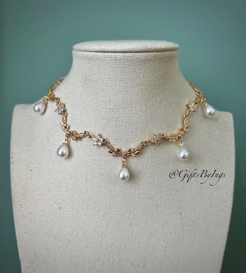 May include: A gold-toned necklace with floral and leaf detailing, adorned with clear crystals and dangling teardrop-shaped pearls. The necklace is displayed on a mannequin head against a teal background. The text "@GiftsByIngs" is visible.