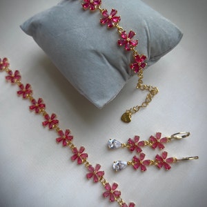 May include: A gold jewelry set with pink flower-shaped charms and clear teardrop-shaped stones. The set includes a bracelet, a pair of earrings, and a necklace.