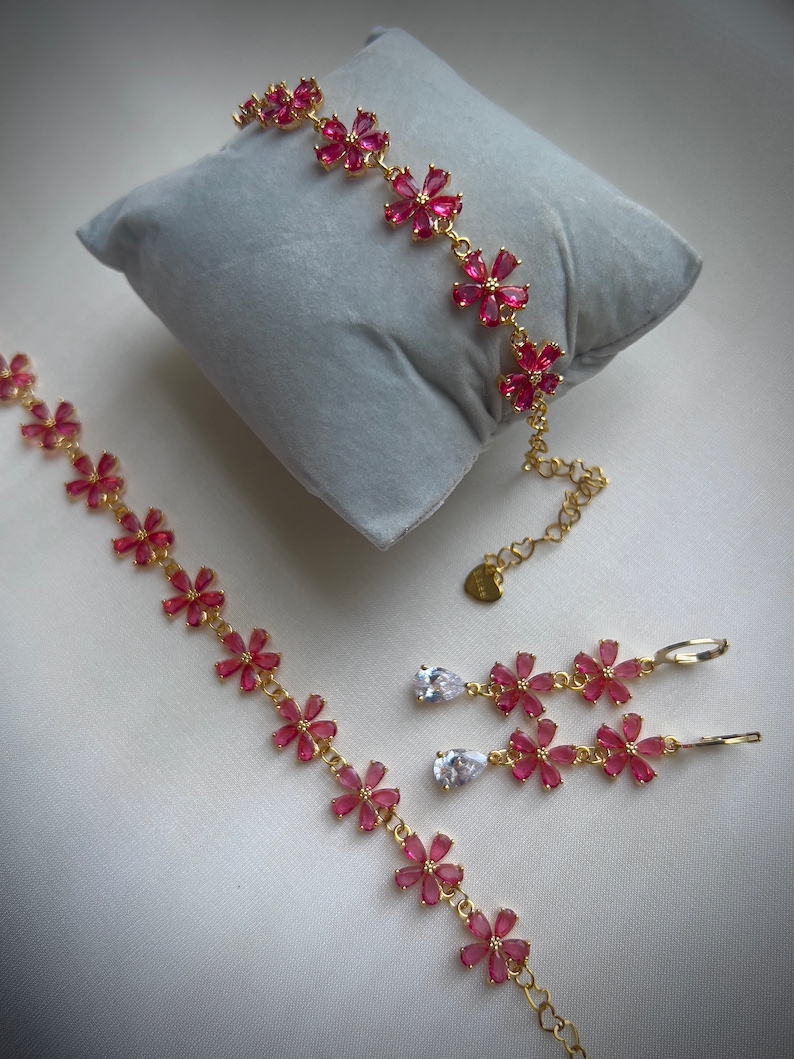 May include: A gold jewelry set with pink flower-shaped charms and clear teardrop-shaped stones. The set includes a bracelet, a pair of earrings, and a necklace.