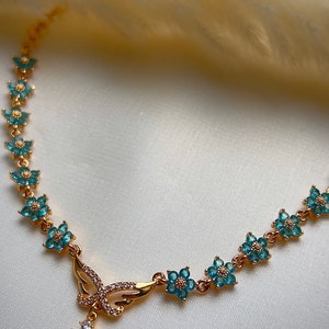 Puede incluir: Un collar de oro con un colgante de mariposa y detalles de flores azules. El colgante tiene cristales transparentes y una piedra ovalada azul claro.