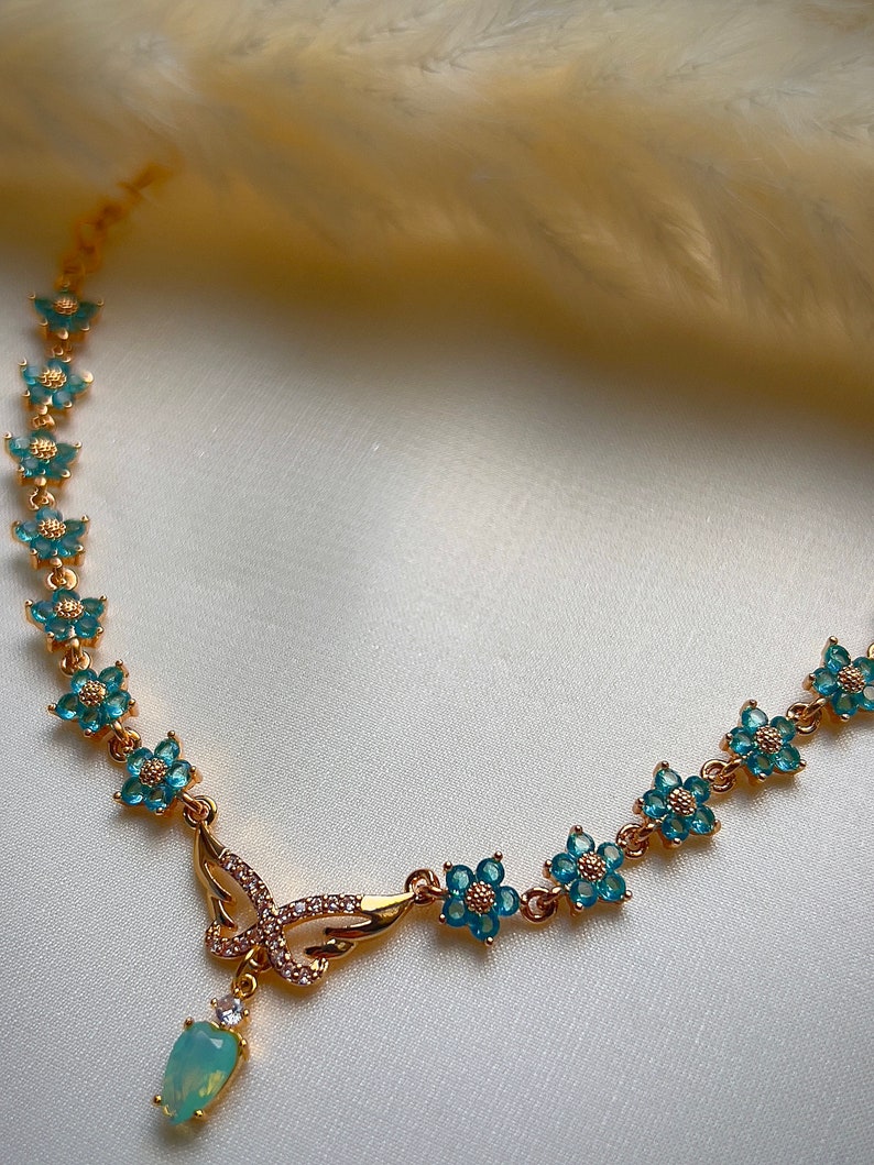Puede incluir: Un collar de oro con un colgante de mariposa y detalles de flores azules. El colgante tiene cristales transparentes y una piedra ovalada azul claro.