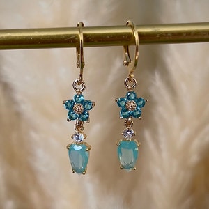 Puede incluir: Pendientes de aro dorados con un dije de flor azul y una piedra azul en forma de l&aacute;grima.
