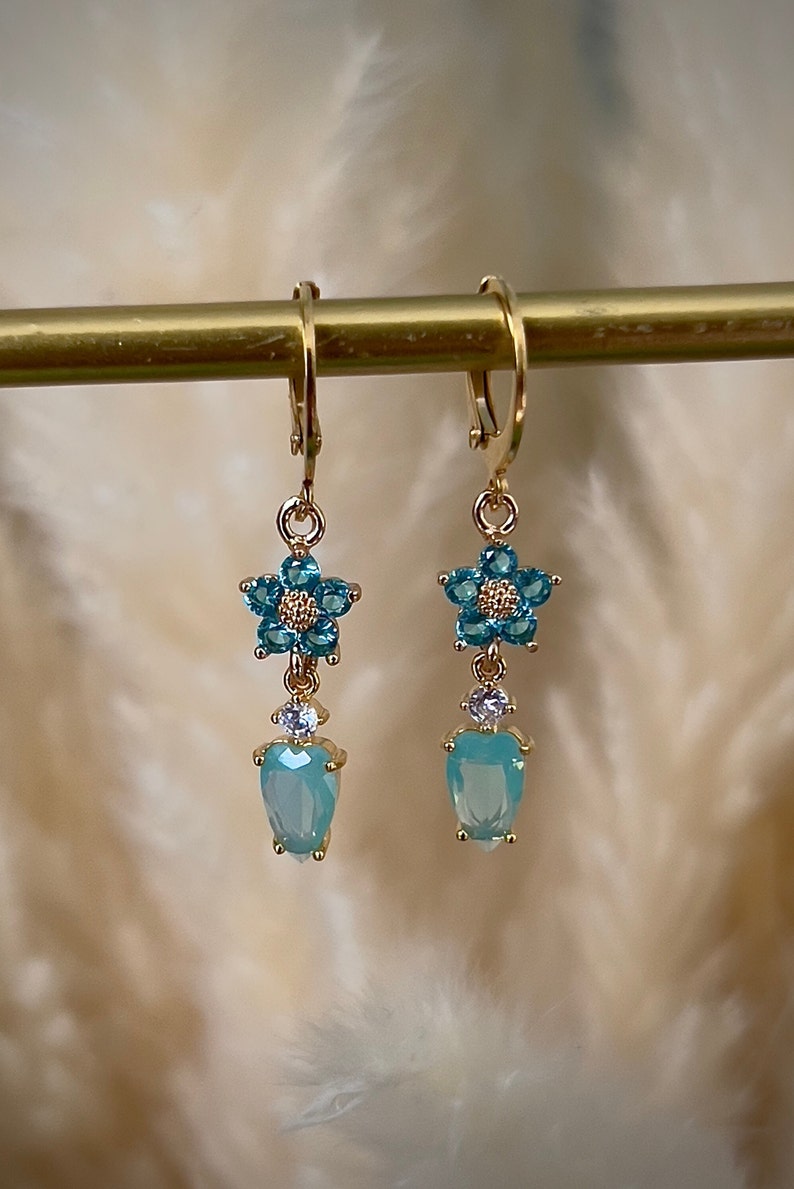 Puede incluir: Pendientes de aro dorados con un dije de flor azul y una piedra azul en forma de l&aacute;grima.