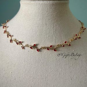 Può includere: Una collana dorata con un delicato design a vite, adornata con piccole foglie di gemme rosse. La collana è esposta su un manichino, mettendo in risalto la sua estetica elegante e ispirata alla natura. Il testo "@GiftsByIngs" è visibile.