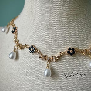 May include: A gold-toned necklace with a floral design, black flower accents, and dangling teardrop-shaped pearls. The necklace is displayed on a mannequin. The necklace has the text "@GiftsByInys".