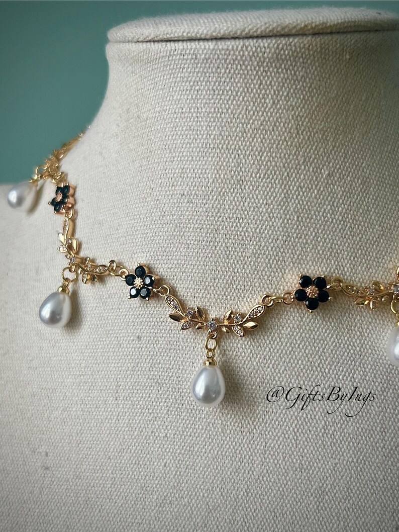 May include: A gold-toned necklace with a floral design, black flower accents, and dangling teardrop-shaped pearls. The necklace is displayed on a mannequin. The necklace has the text "@GiftsByInys".