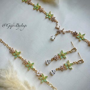 May include: A gold-toned jewelry set featuring a necklace and matching drop earrings. The pieces are adorned with light green flower-shaped stones and clear crystals. The necklace includes a central teardrop crystal. The text @GiftsByIngs is visible.