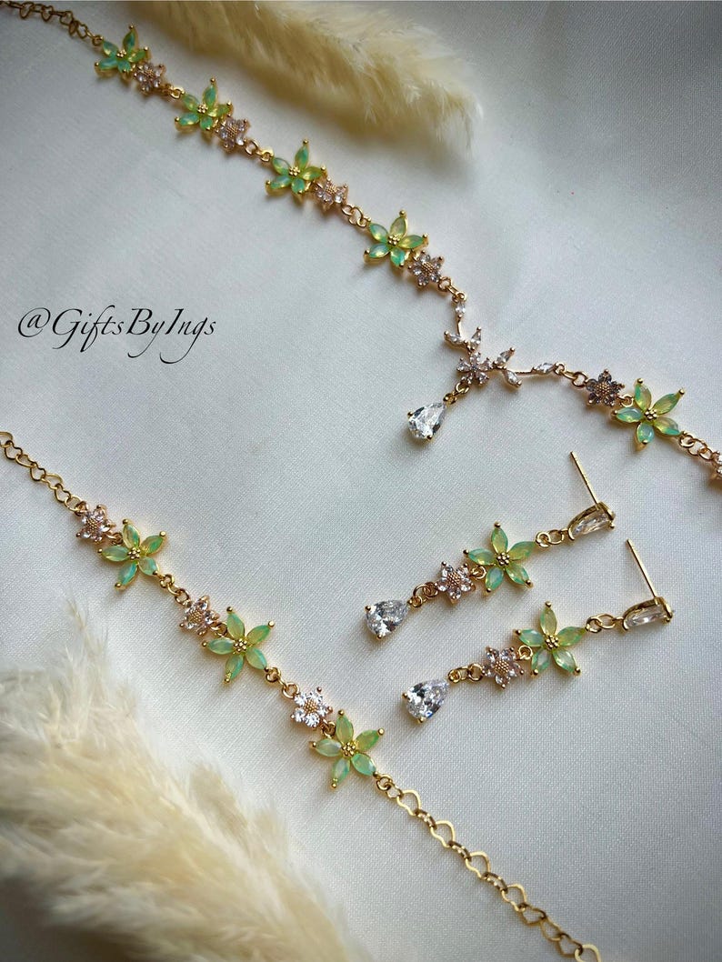 May include: A gold-toned jewelry set featuring a necklace and matching drop earrings. The pieces are adorned with light green flower-shaped stones and clear crystals. The necklace includes a central teardrop crystal. The text @GiftsByIngs is visible.