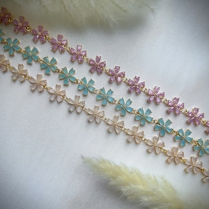 May include: Three gold chains with delicate flower charms. The flowers are in shades of pink, light blue, and white.