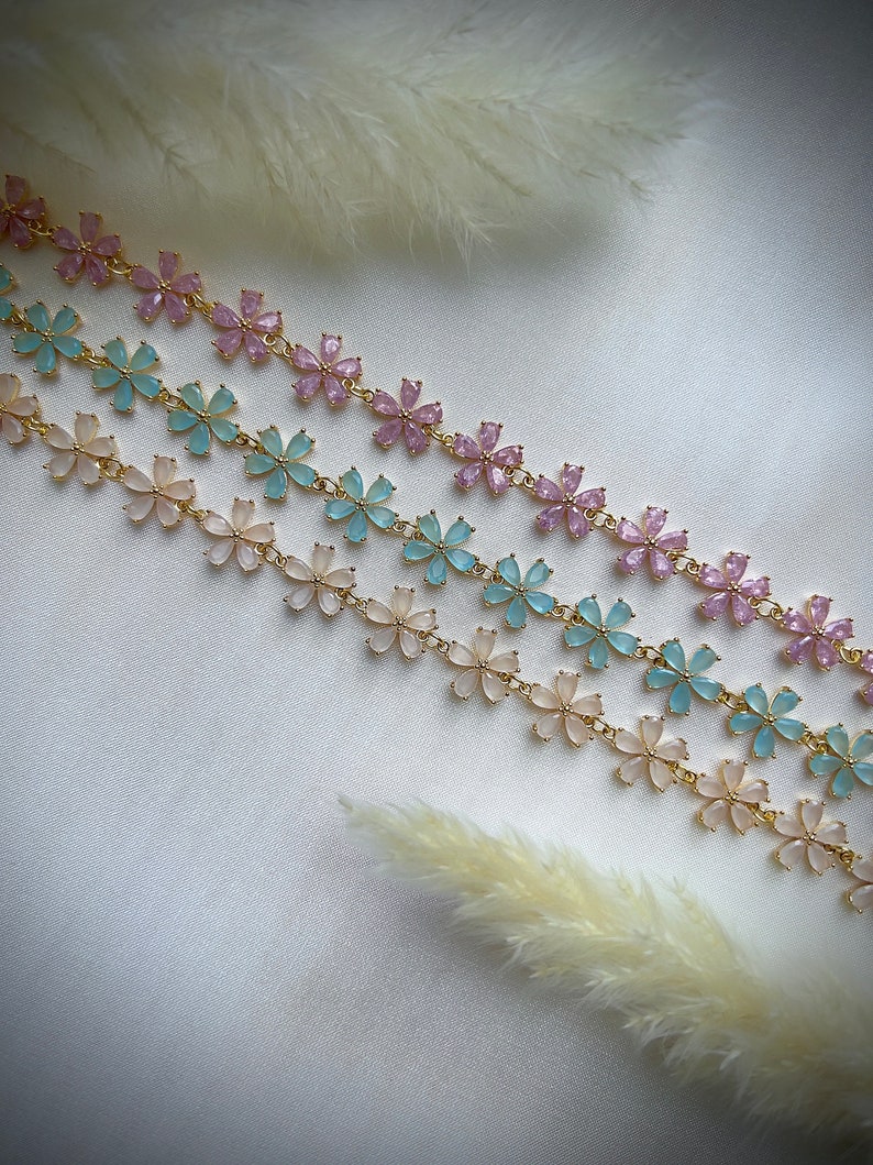 May include: Three gold chains with delicate flower charms. The flowers are in shades of pink, light blue, and white.