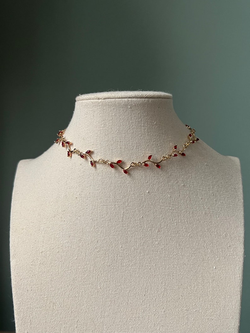 May include: A gold chain necklace with a delicate vine design and red gemstone accents.