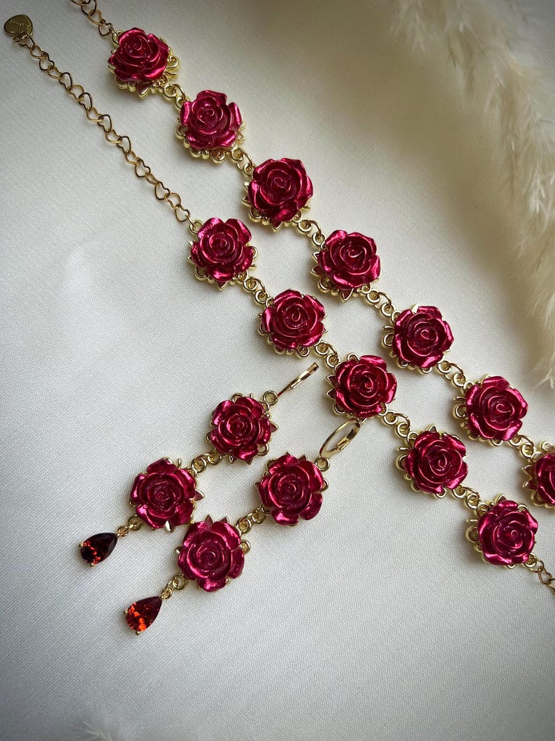 May include: A set of rose-shaped jewelry featuring a pair of earrings and a bracelet. The earrings have three rose-shaped charms with red gemstone accents. The bracelet has a gold chain with multiple rose-shaped charms.