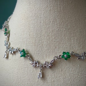 May include: A silver necklace with a delicate design featuring green and clear crystal flowers and leaves. The necklace has a dangling crystal drop.