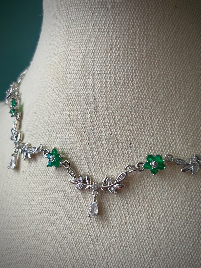 May include: A silver necklace with a delicate design featuring green and clear crystal flowers and leaves. The necklace has a dangling crystal drop.