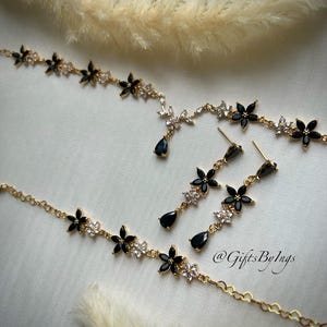 May include: A gold-toned jewelry set consisting of a necklace and matching drop earrings. The pieces feature alternating black and clear crystal flower motifs, with the necklace showcasing a central teardrop pendant and the earrings featuring a teardrop drop. The text @GiftsByIngs is visible.