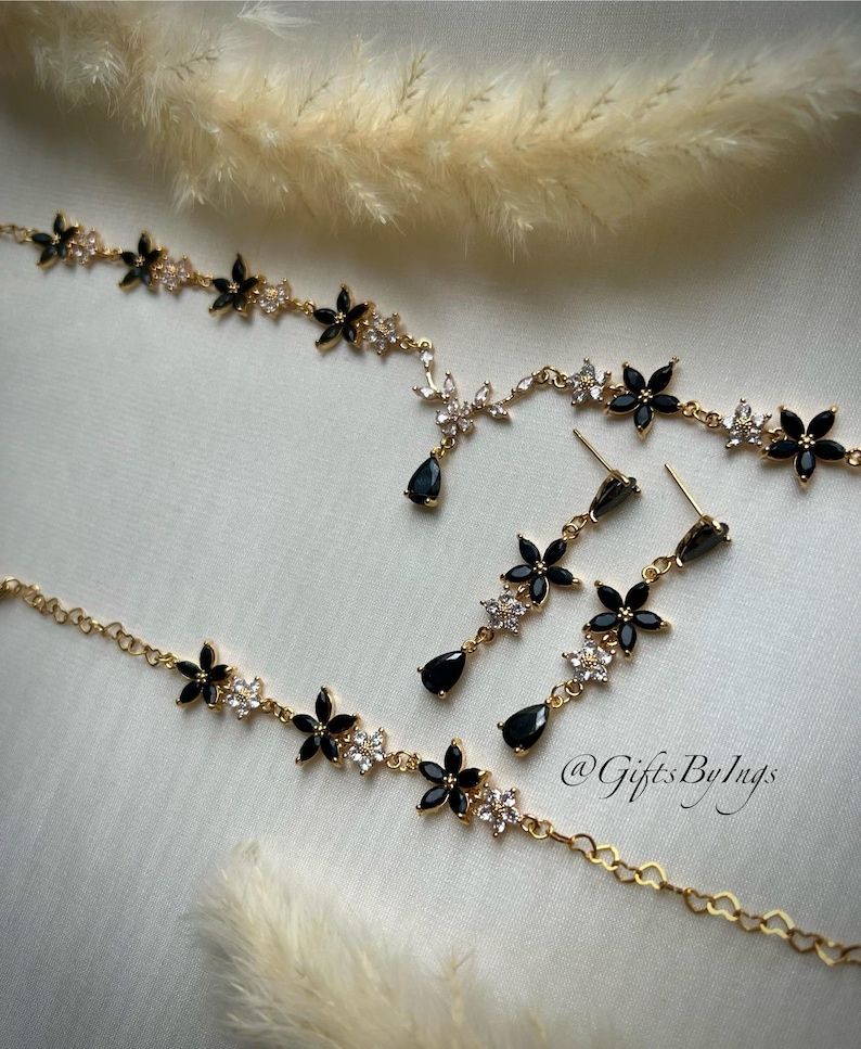 May include: A gold-toned jewelry set consisting of a necklace and matching drop earrings. The pieces feature alternating black and clear crystal flower motifs, with the necklace showcasing a central teardrop pendant and the earrings featuring a teardrop drop. The text @GiftsByIngs is visible.