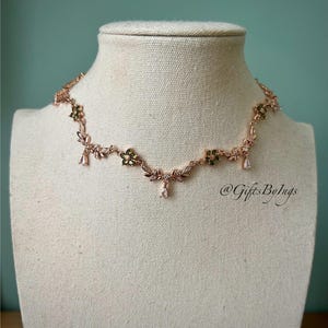 Puede incluir: Collar de oro rosa con diseño floral y de hojas. El collar presenta pequeñas piedras preciosas verdes y transparentes, con colgantes en forma de lágrima. El collar se exhibe en un maniquí.
