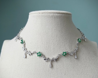 May include: A silver necklace with a delicate floral design featuring green gemstones. The necklace has a pendant with a teardrop-shaped crystal.