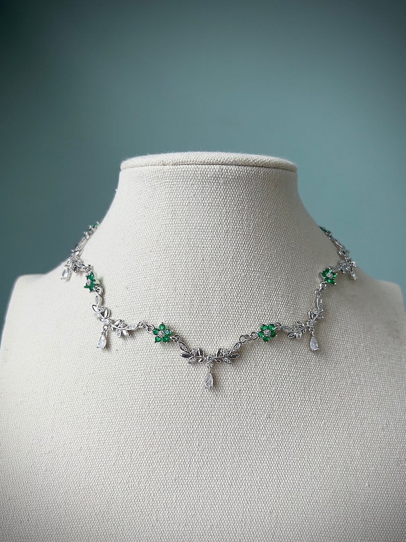May include: A silver necklace with a delicate floral design featuring green gemstones. The necklace has a pendant with a teardrop-shaped crystal.