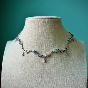 May include: A silver necklace with a delicate floral design and blue gemstones. The necklace features a pendant with a teardrop-shaped gemstone.