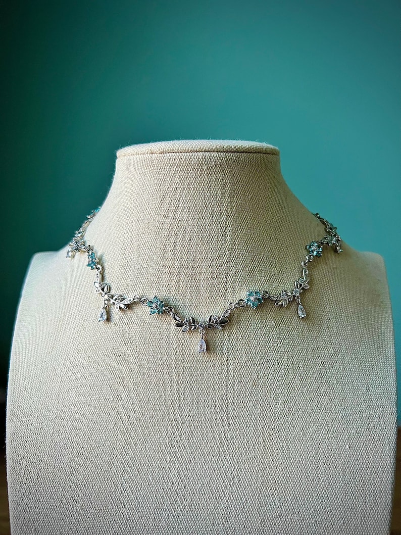 May include: A silver necklace with a delicate floral design and blue gemstones. The necklace features a pendant with a teardrop-shaped gemstone.