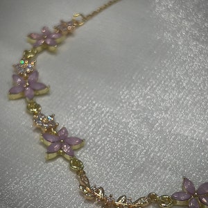 May include: A gold chain necklace with a delicate floral design. The necklace features pink flowers with clear rhinestones and a dangling crystal charm.