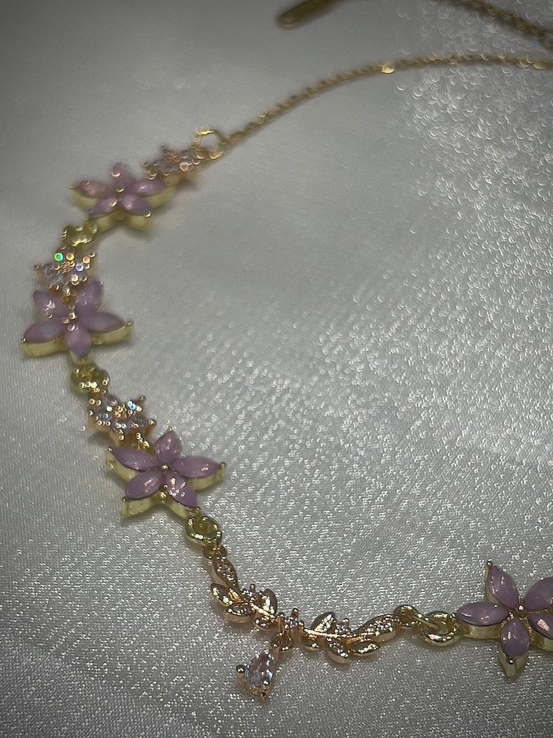 May include: A gold chain necklace with a delicate floral design. The necklace features pink flowers with clear rhinestones and a dangling crystal charm.