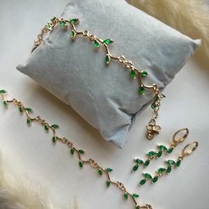 May include: A gold necklace, bracelet, and earrings set with green gemstone leaves. The jewelry is delicate and feminine.