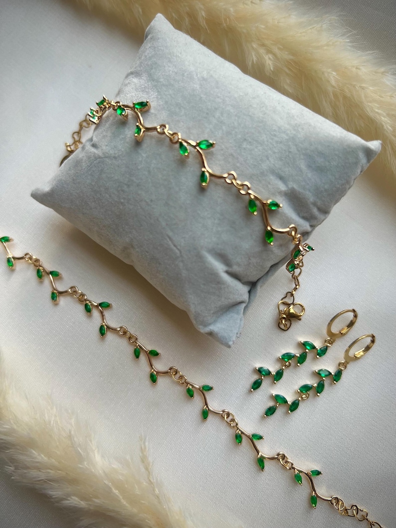 May include: A gold necklace, bracelet, and earrings set with green gemstone leaves. The jewelry is delicate and feminine.