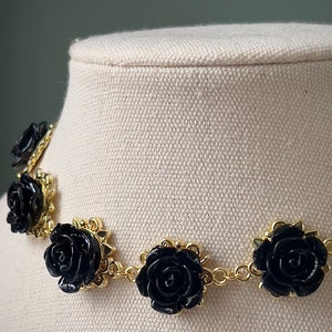 May include: A black rose choker necklace with gold chain links and black rose accents. The necklace is on a white fabric background.
