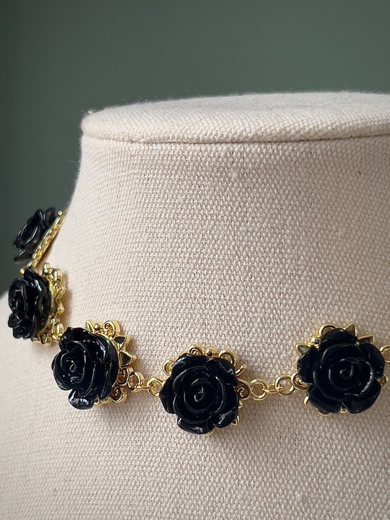 May include: A black rose choker necklace with gold chain links and black rose accents. The necklace is on a white fabric background.