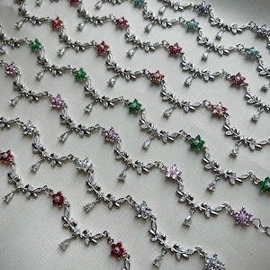 May include: A collection of silver anklets with delicate floral designs and dangling charms. Each anklet features a different colored gemstone in the center of the flower.