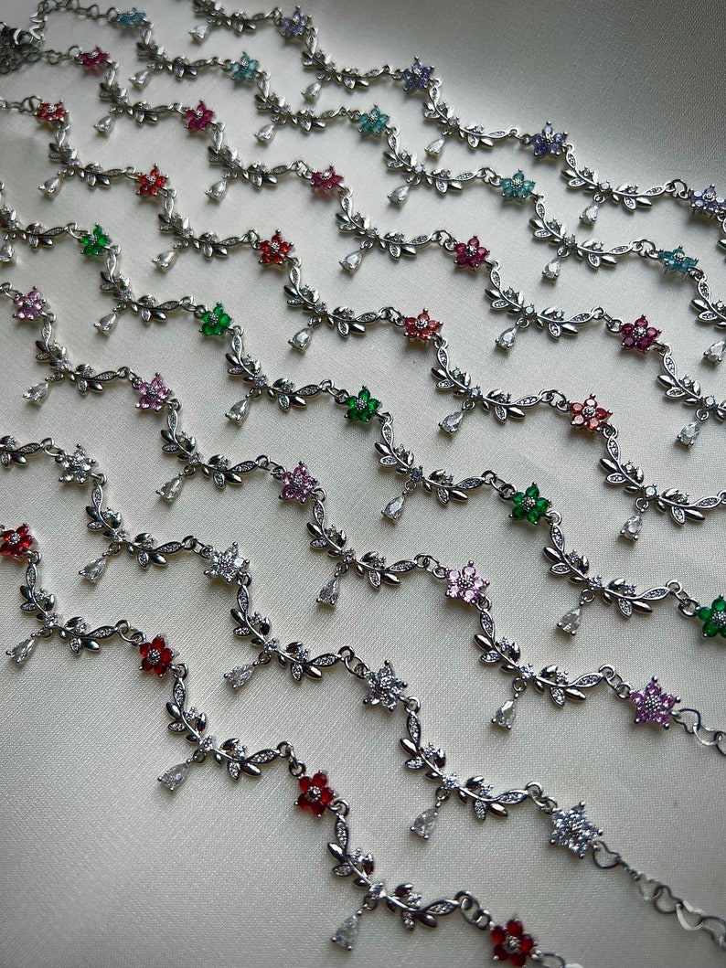 May include: A collection of silver anklets with delicate floral designs and dangling charms. Each anklet features a different colored gemstone in the center of the flower.