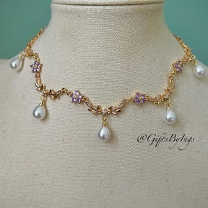 May include: A gold-toned necklace with a floral design, featuring purple flower accents and dangling teardrop-shaped pearls. The necklace is displayed on a mannequin. The text "@GiftsByIngs" is visible.