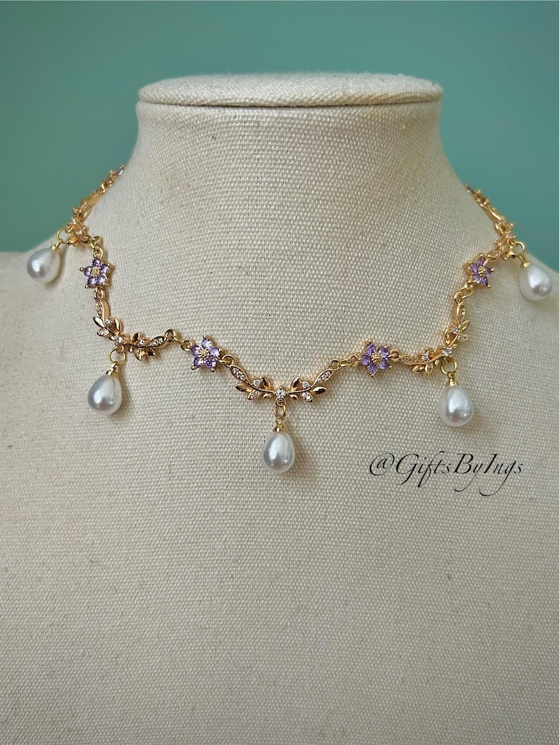 May include: A gold-toned necklace with a floral design, featuring purple flower accents and dangling teardrop-shaped pearls. The necklace is displayed on a mannequin. The text "@GiftsByIngs" is visible.