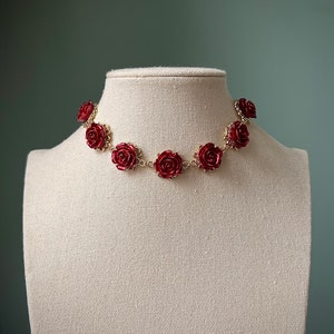 May include: A gold chain necklace with red rose-shaped charms. The necklace is displayed on a white mannequin.