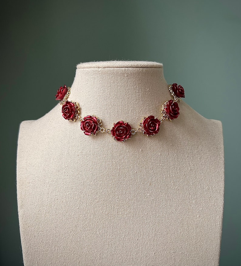 May include: A gold chain necklace with red rose-shaped charms. The necklace is displayed on a white mannequin.