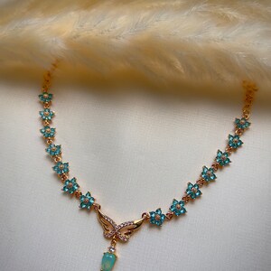 Puede incluir: Un collar de oro con un colgante de mariposa y detalles de flores azules. El colgante tiene una piedra ovalada azul claro.