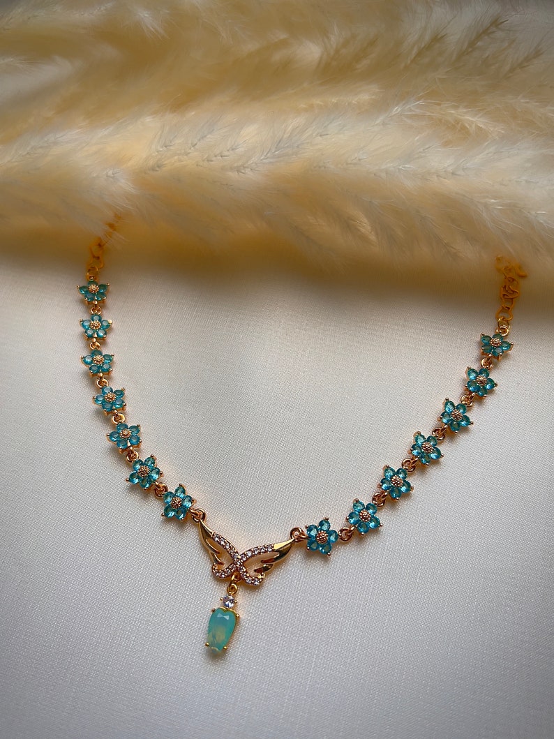 Puede incluir: Un collar de oro con un colgante de mariposa y detalles de flores azules. El colgante tiene una piedra ovalada azul claro.