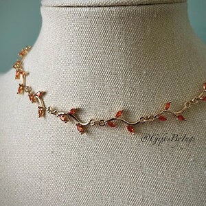 May include: A gold necklace with a vine design and orange gemstones. The necklace is on a white mannequin.