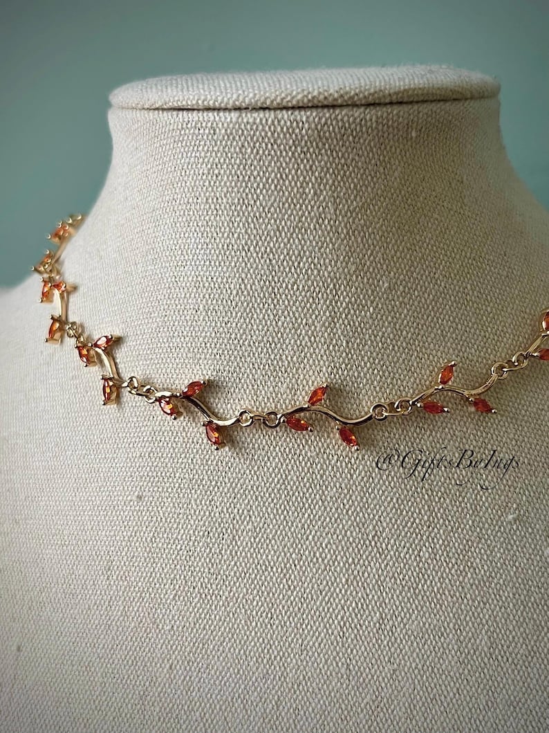 May include: A gold necklace with a vine design and orange gemstones. The necklace is on a white mannequin.